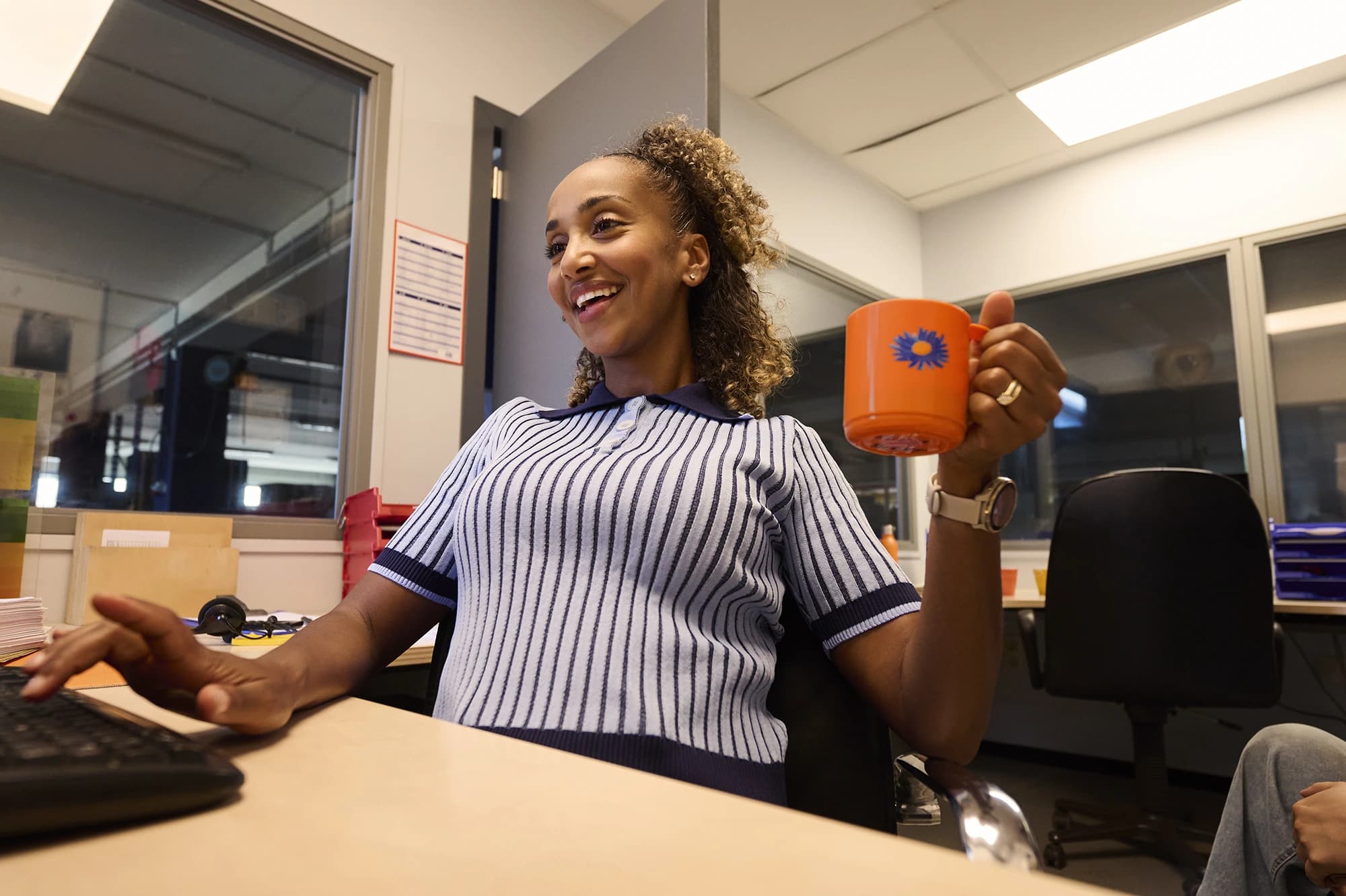 Colaborador administrativo sorri com uma caneca de café atrás do computador — empregos de escritório com prazer no trabalho via Accent